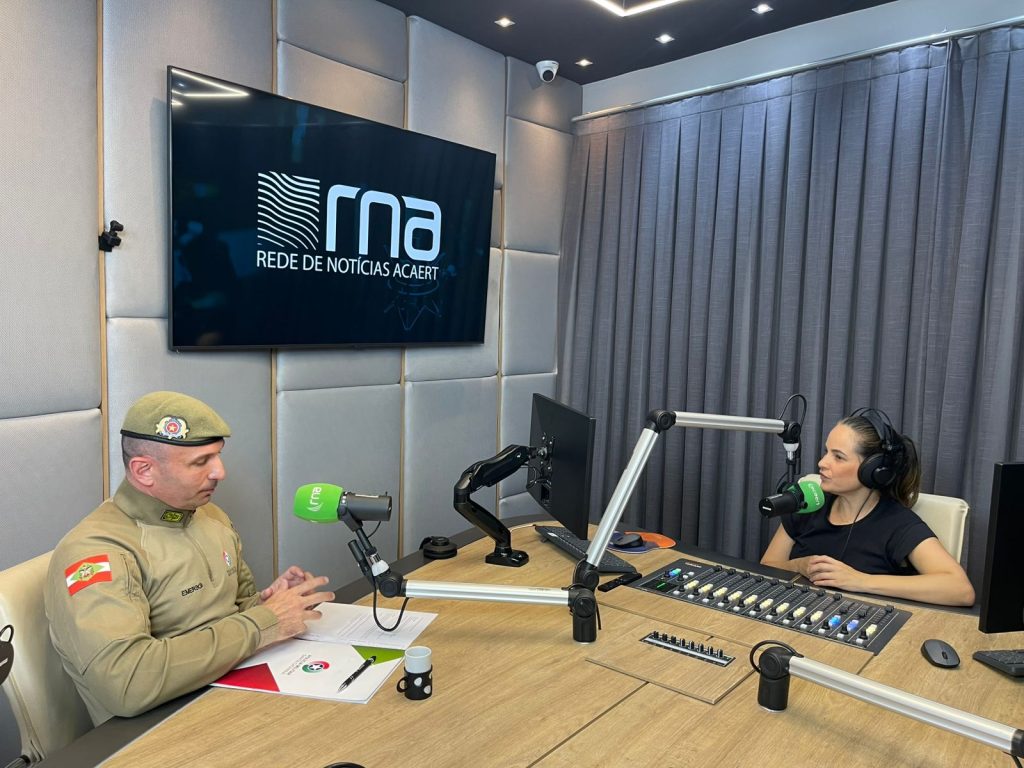 Entrevista em estúdio de rádio: oficial da Polícia Militar, fardado, conversa ao microfone com apresentadora, em ambiente profissional da RNA/ACAERT.
