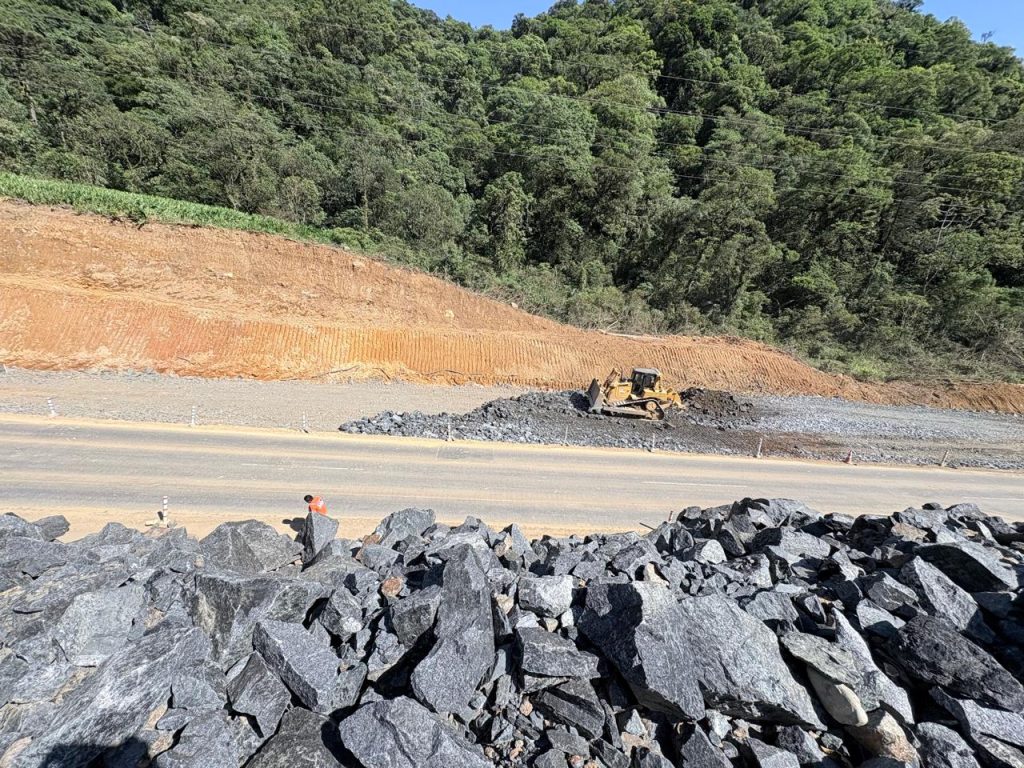 Obras de área de escape na SC-418 mostram terraplenagem, máquinas e material de contenção em trecho da Serra Dona Francisca, com pista ao fundo e vegetação nas encostas.