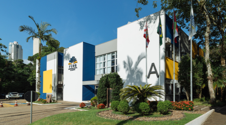 Prédio da Fundação Universidade Regional de Blumenau (FURB) com fachada moderna, jardins e bandeiras institucionais.