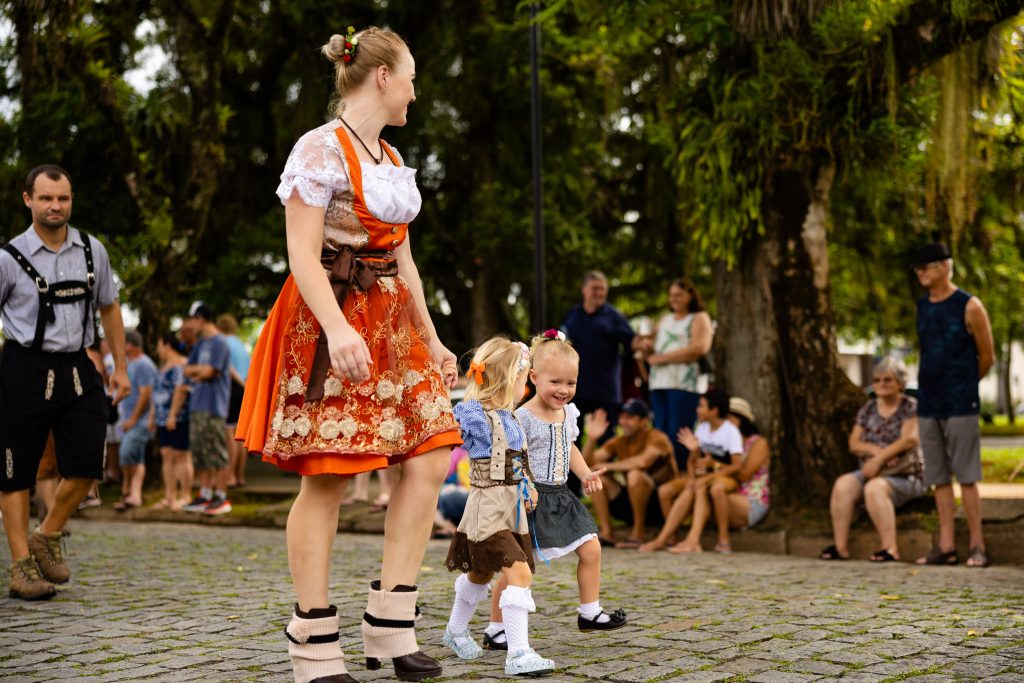 Mulher e duas crianças caminham em desfile usando trajes típicos alemães durante a Festa Pomerana, em Pomerode, com público ao fundo