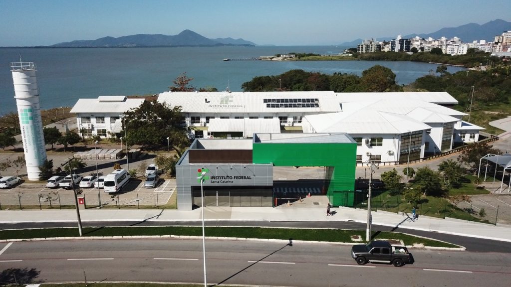 Vista aérea do câmpus do Instituto Federal de Santa Catarina, com prédios administrativos e acadêmicos, estacionamento, via urbana e área costeira ao fundo.