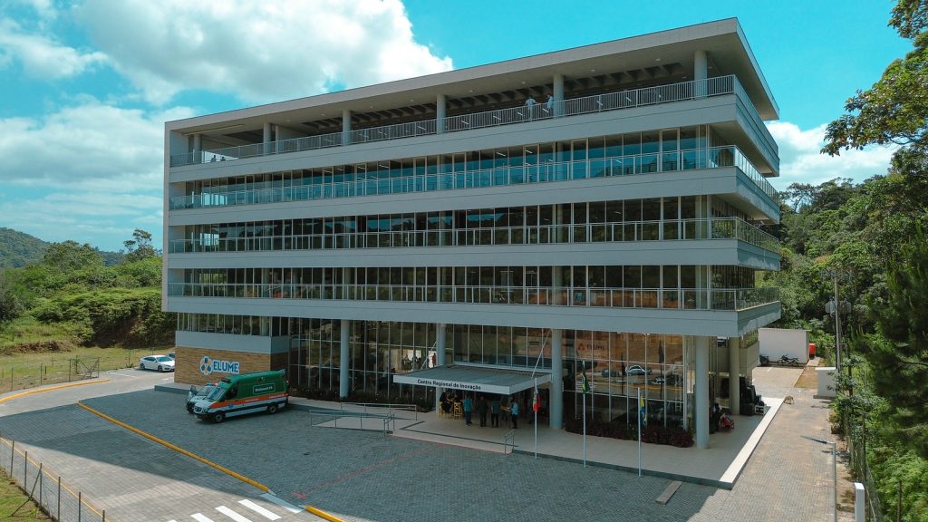 Fachada de Centro de Inovação em Santa Catarina, com prédio moderno, área externa arborizada e veículos estacionados.
