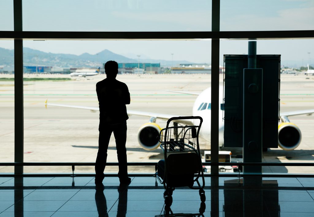 Passageiro em pé dentro de aeroporto observa aeronave estacionada na pista por meio de uma parede de vidro.