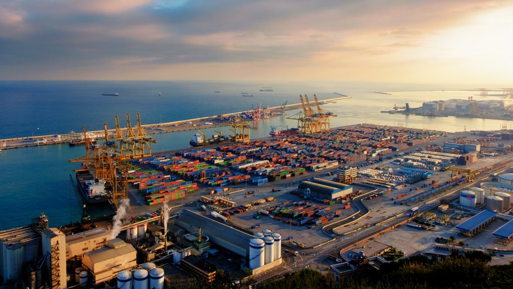Vista aérea de um porto com contêineres coloridos empilhados, guindastes e navios cargueiros, representando a movimentação do comércio exterior.
