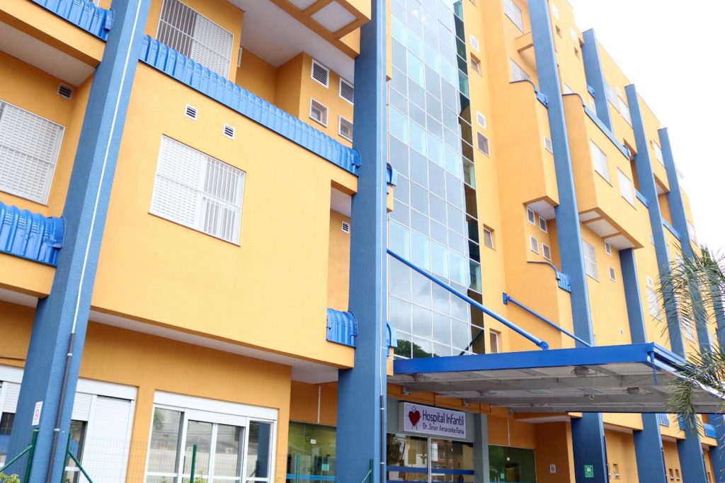 Fachada do Hospital Infantil Dr. Jeser Amarante Faria, em Joinville, com prédio nas cores amarelo e azul, janelas, varandas e entrada principal coberta.