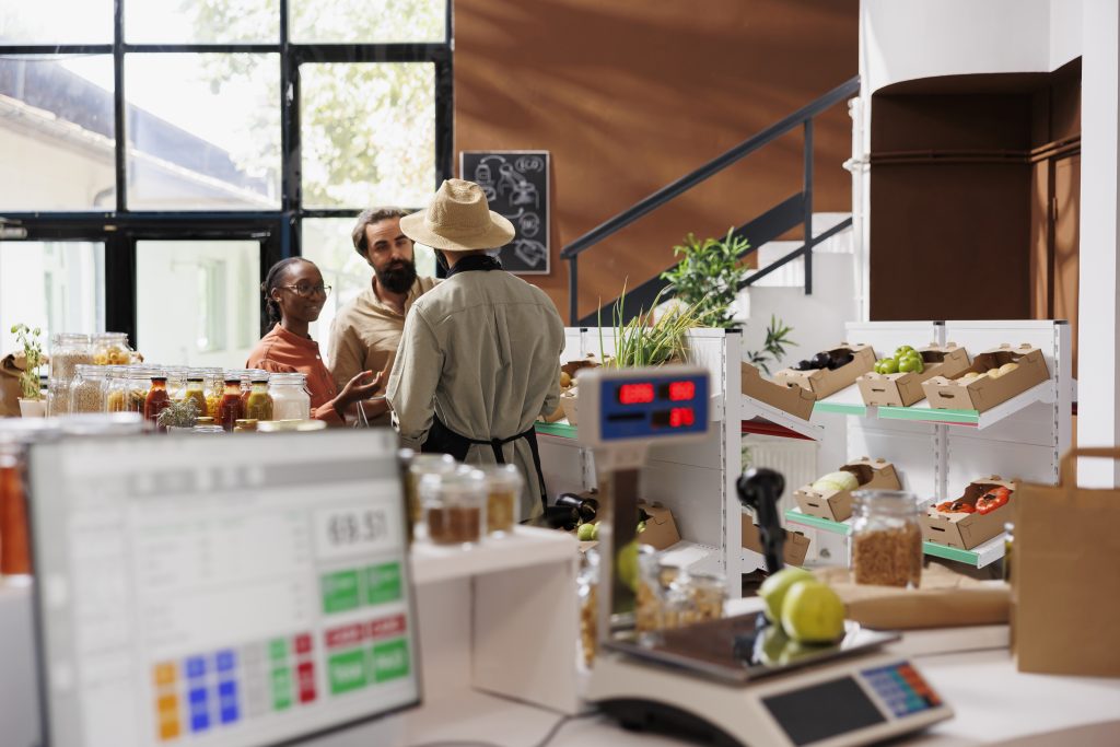 Pessoas atendendo clientes em um pequeno comércio, com produtos expostos em prateleiras e balanças no balcão, representando a rotina de uma microempresa.